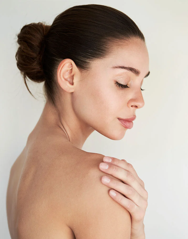 A profile shot of a woman with dark hair pulled back in a bun, her eyes closed and head tilted slightly downward. She is bare-shouldered, and her left hand is gently resting on her right shoulder. The background is a plain, light color. - Arm Lift in Folsom, CA