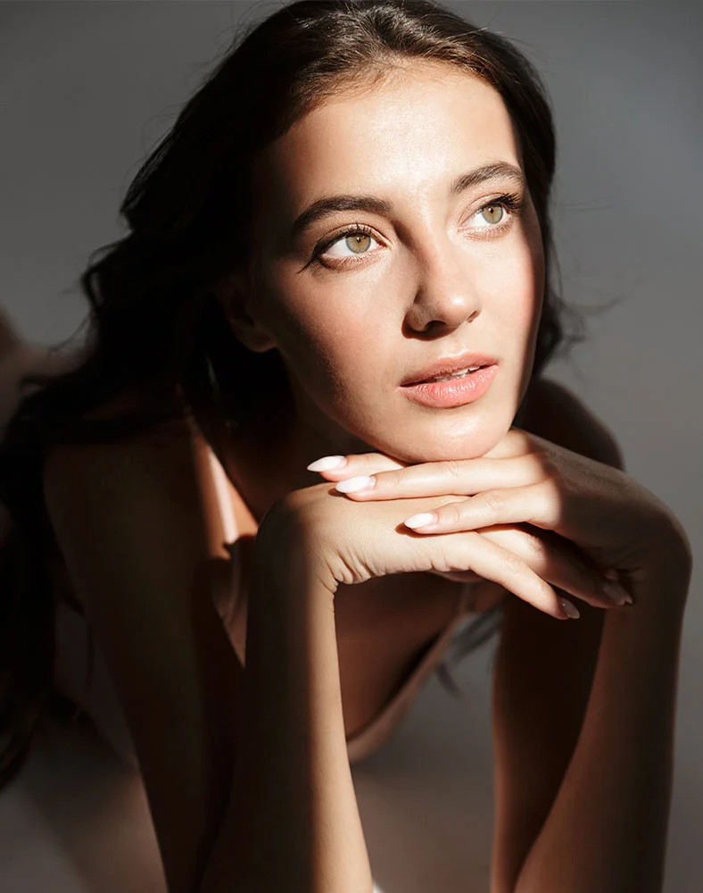 A close-up image of a woman with long, dark hair and light-colored eyes. She is lying on her stomach, resting her chin on her hands, and looking upward to the side. A ray of sunlight shines diagonally across her face and body. - Chemical Peels in Folsom, CA