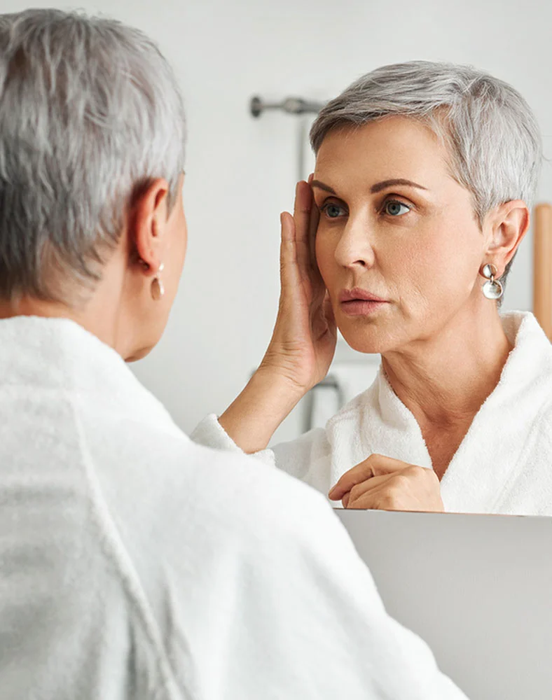 A woman with short, gray hair is looking at her reflection in a mirror. She is wearing a white bathrobe. She is touching her face, and her expression appears somewhat concerned or pensive. The lighting in the bathroom is bright and neutral. The background is a light gray/white bathroom wall. The reflection in the mirror shows a similar-aged woman with a similar expression. - Eyelid Surgery in Folsom, CA