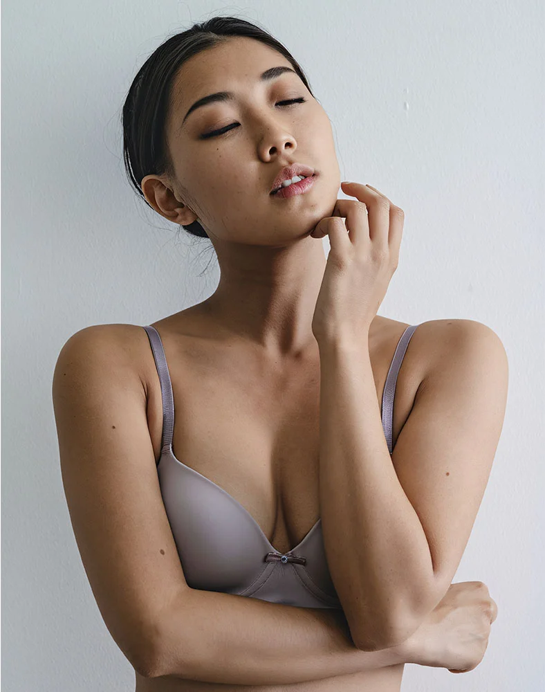 A woman with her eyes closed is standing in front of a white wall, touching her chin and neck with her right hand. She is wearing a simple gray bra with a small bow in the center. - Fat Grafting Breast Augmentation in Folsom, CA