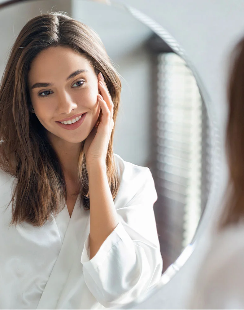 A young woman with shoulder-length brown hair is looking at herself in a round mirror. She is wearing a white robe-like top, and her hand is gently resting on her cheek. Her expression is pleasant and she is smiling. The background is out of focus, showing a blurred light-colored interior, likely a bathroom or bedroom. The lighting is soft and natural. - Laser Resurfacing in Folsom, CA