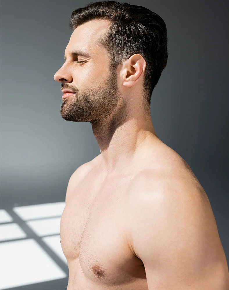 A slightly high-angle, close-up profile view of a light-skinned man with short dark brown hair and a well-groomed beard. His eyes are closed, and he appears relaxed. His toned chest and shoulders are visible. Soft, diffused light highlights his features, while a window grid shadow on the floor adds depth to the muted gray background. - Male Breast Reduction Gynecomastia in Folsom, CA