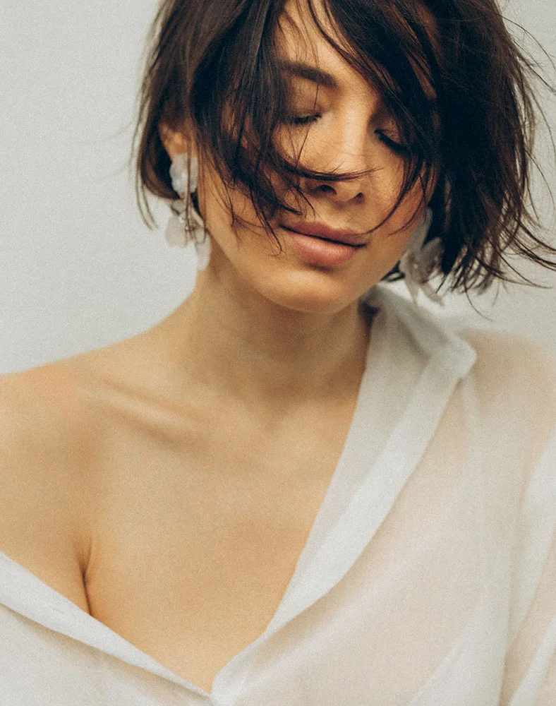A woman with short, tousled dark brown hair is shown from the mid-chest up, wearing a loose, off-white button-down shirt with the top buttons undone, exposing her shoulders. Her eyes are closed and her expression is calm. The background is a plain light beige or gray, softly lit to create an ethereal feel. - Neck Lift in Folsom, CA