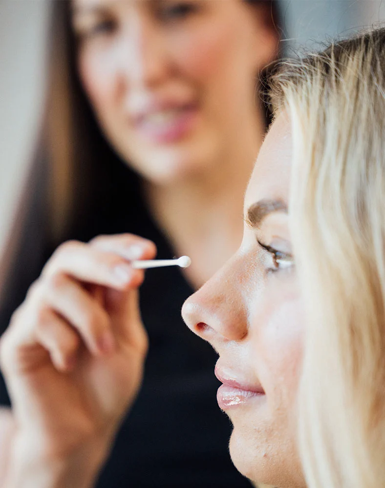 Close-up of Dr. Daly, a makeup artist with dark hair, applying makeup near the bridge of a light blonde woman’s nose using a cotton swab or small applicator. The woman’s eyes are open with a neutral expression and light pinkish-nude lips. Soft lighting highlights her smooth skin, while the blurred background keeps focus on her face and Dr. Daly’s hands. - Rhinoplasty in Folsom, CA